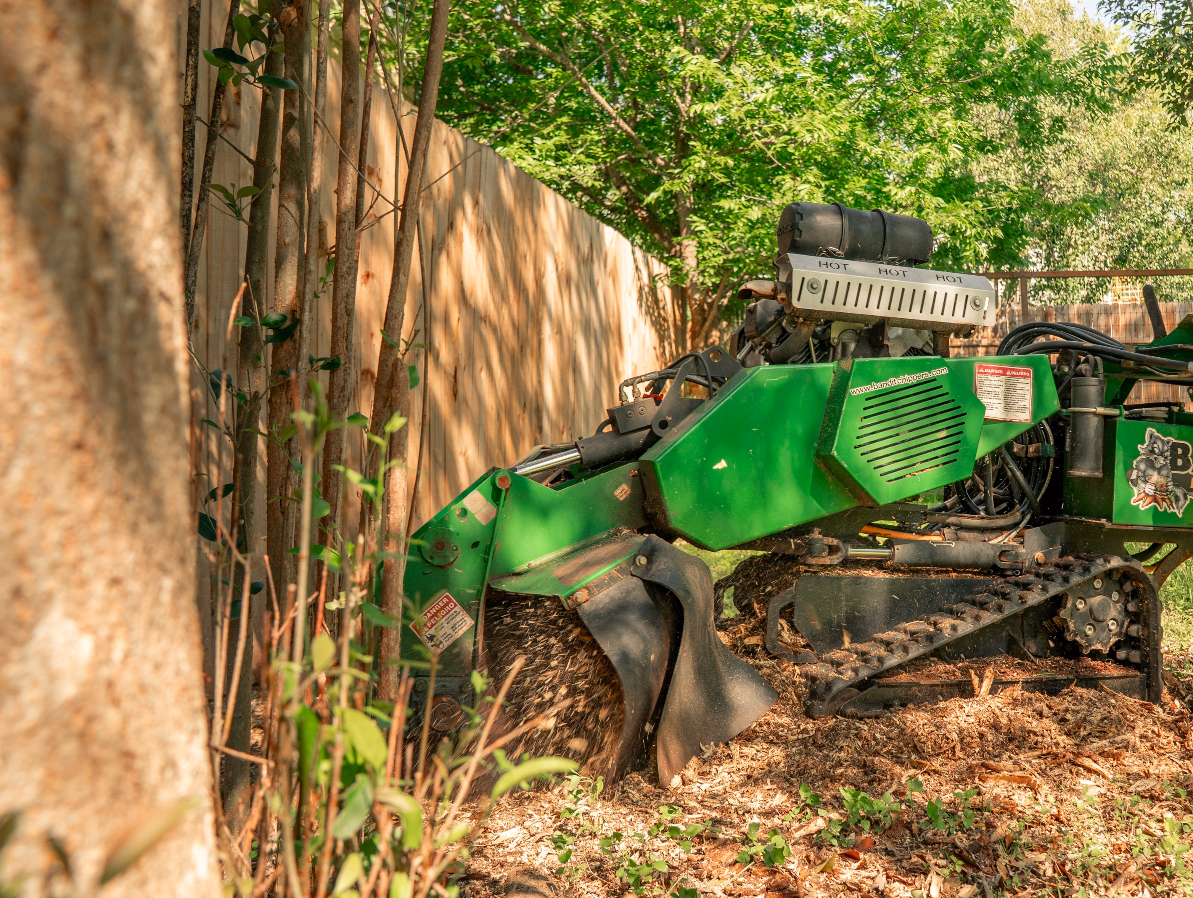 Green stump grinder cutting a tree stump near a wooden fence with wood chips flying.